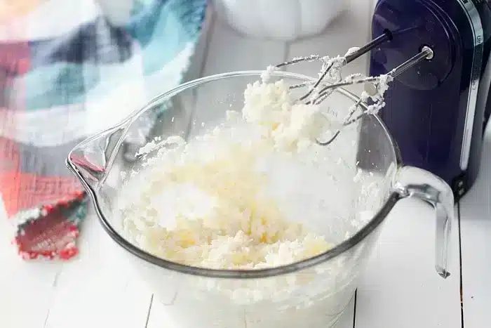 A clear glass mixing bowl containing a creamy mixture is placed on a white surface. An electric hand mixer with beaters covered in the mixture rests in the bowl, ready to whip up a 1/2 sheet cake.