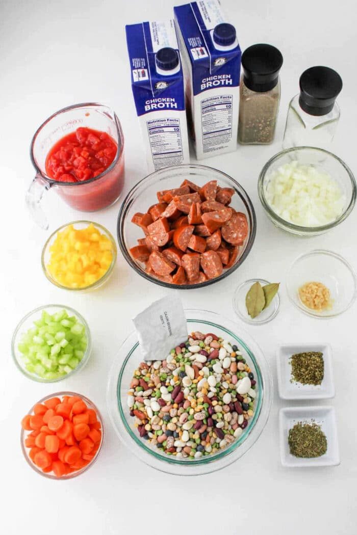 Assorted 15 Bean Soup Crock Pot ingredients in bowls: beans, sausage, vegetables, canned tomatoes, broth, and spices on a white background.