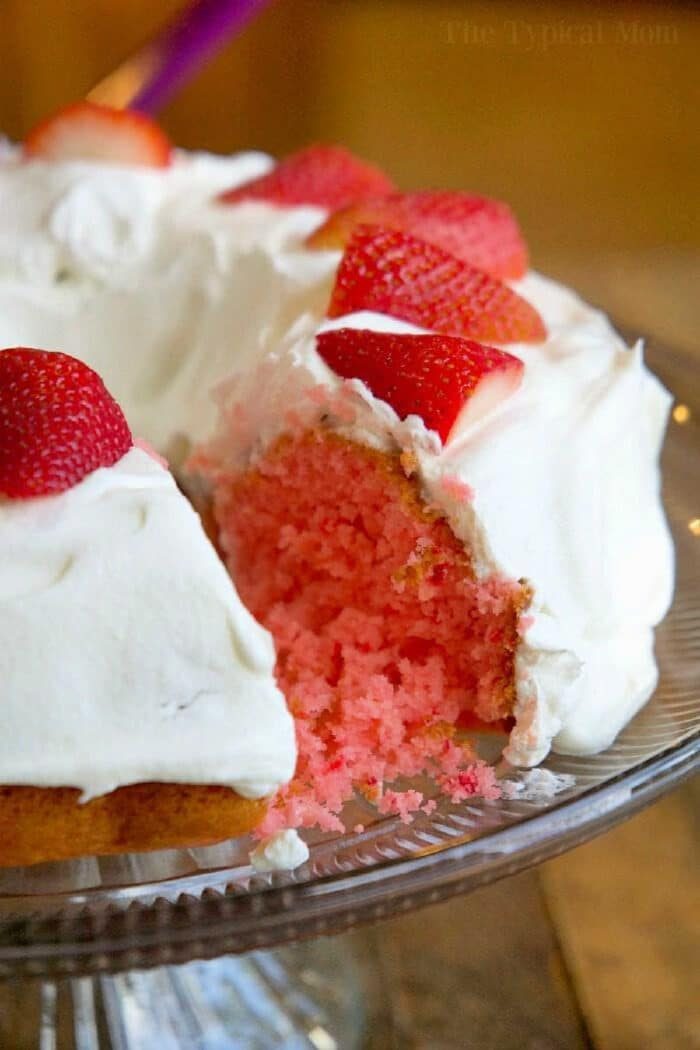 The delightful 2 ingredient strawberry cake features white frosting and is adorned with sliced strawberries on top. Presented elegantly on a glass platter, it tempts with a piece already missing.