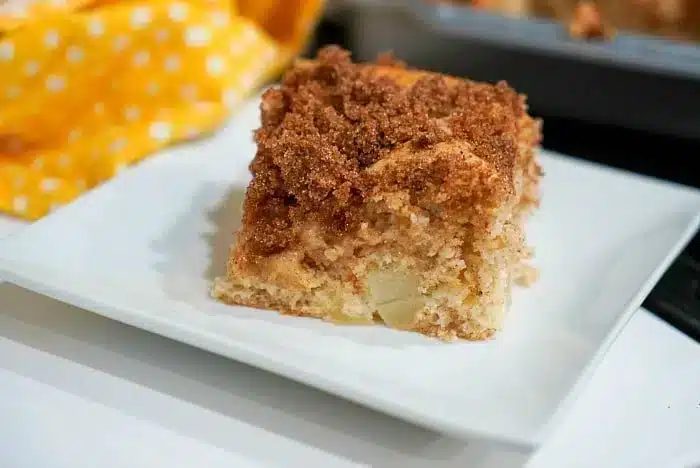 A square slice of crumb-topped coffee cake sits on a white plate. The cake appears moist with a golden brown crumb coating, reminiscent of a simple 3 ingredient dump cake.