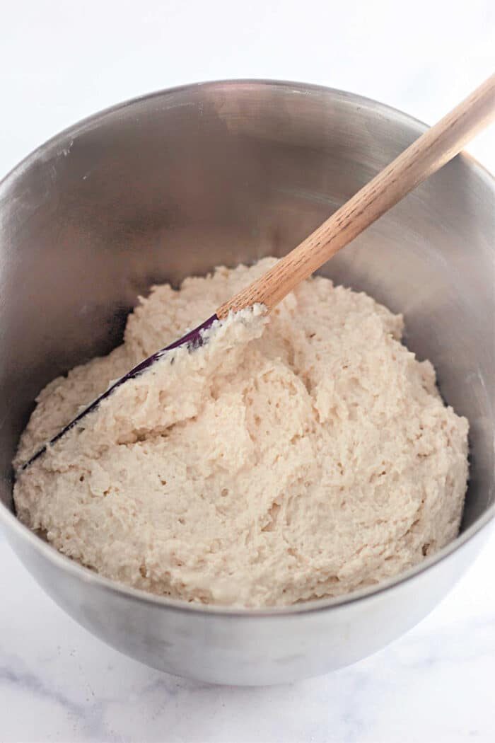A mixing bowl filled with bread dough and a wooden spoon set beside it on a pristine white surface fuses simplicity and charm, reminiscent of crafting 4 Ingredients Basic Biscuits from scratch.