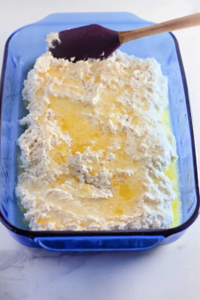 Blue baking dish with bisquick biscuit batter and melted butter, being spread with a spatula to create easy 4 Ingredients Basic Biscuits.