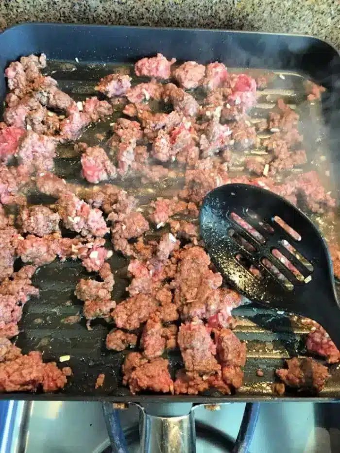 Ground meat sizzles in a grill pan on the stovetop, steam rising as it browns. A slotted black spatula rests atop some of the succulent bits, reminiscent of a 5 ingredient crockpot chili recipe simmering to perfection.