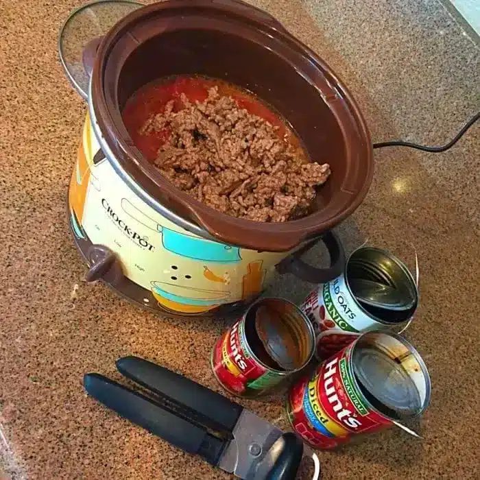 A 5 ingredient crockpot chili recipe sits on a countertop, with a slow cooker containing browned ground meat and tomato sauce.