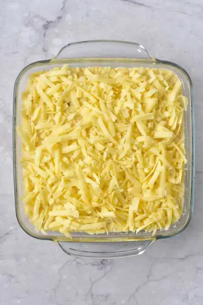 A glass baking dish filled with shredded cheese, placed on a marble countertop. The cheese is spread evenly throughout the dish.