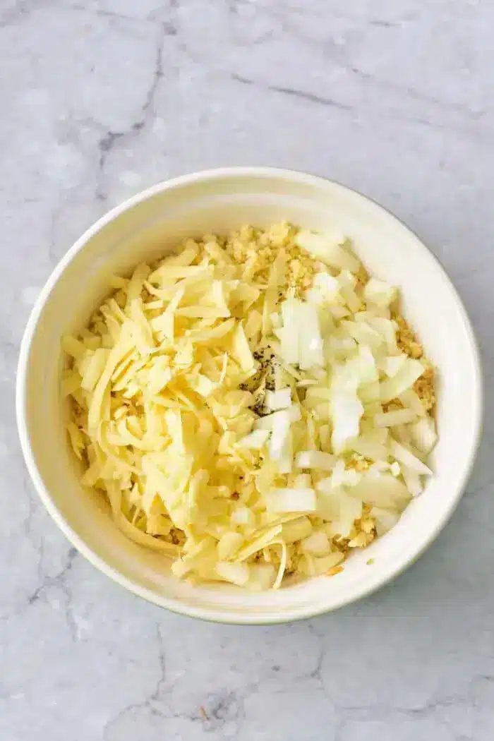 A white bowl filled with shredded cheese, chopped onions, and other finely chopped ingredients, solid for making a good 5 Ingredient Hash Brown Casserole, placed on a marble surface.