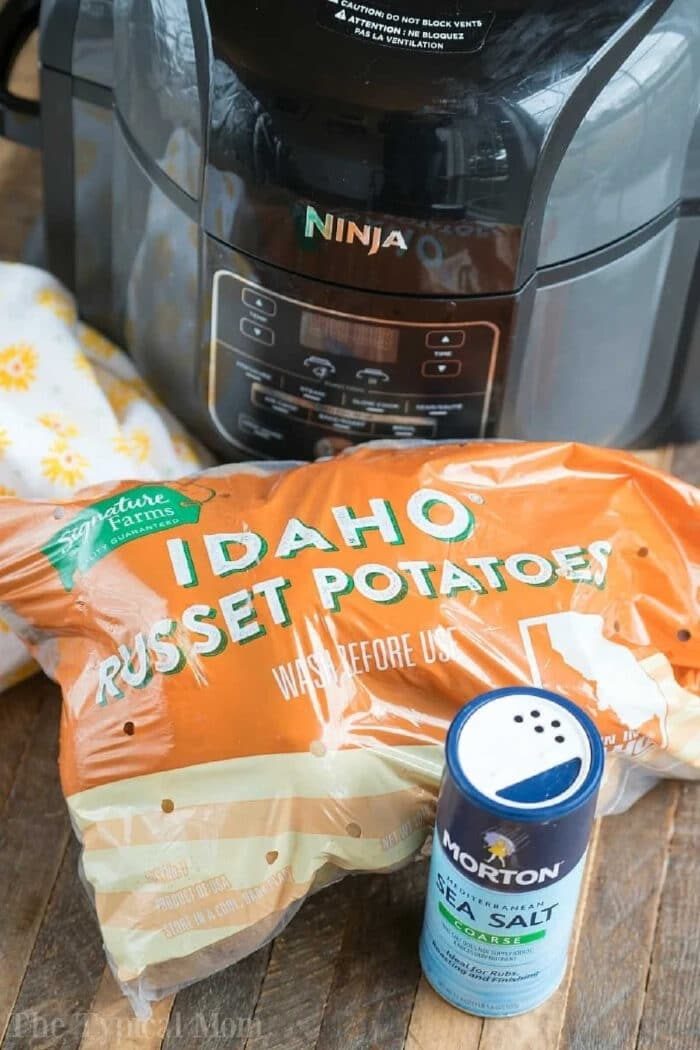 A Ninja air fryer, a bag of Idaho russet potatoes, and a container of sea salt on a wooden surface—everything you need for solid air fryer baked potatoes.
