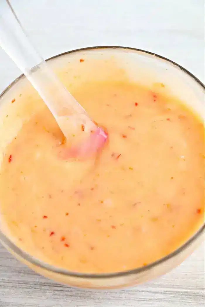 A clear bowl filled with a creamy bang bang sauce containing red flecks, reminiscent of the solid dip for air fryer bang bang shrimp. A transparent spoon is partially submerged in the sauce, and the background is a white surface.