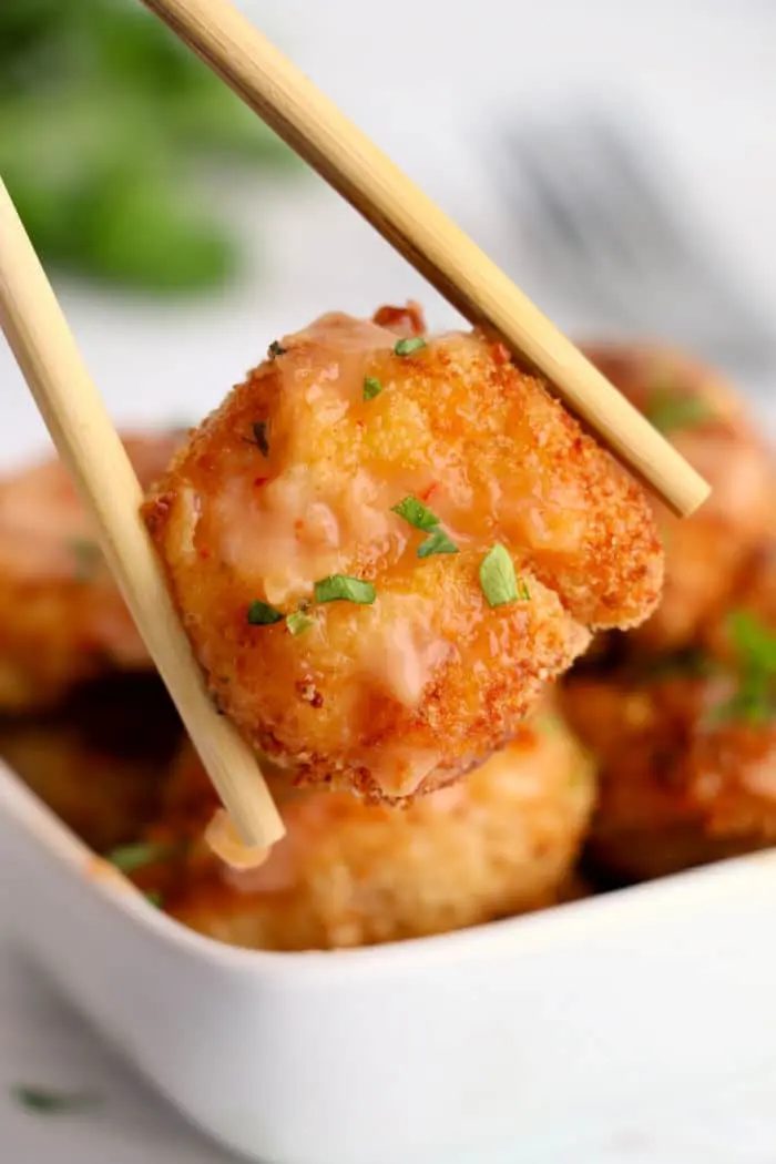 A close-up of chopsticks holding a piece of breaded and fried shrimp, garnished with chopped herbs and a light sauce that gives it an almost bang bang shrimp vibe. In the background, there is a square white bowl filled with more air-fried pieces of the same good breaded tofu.