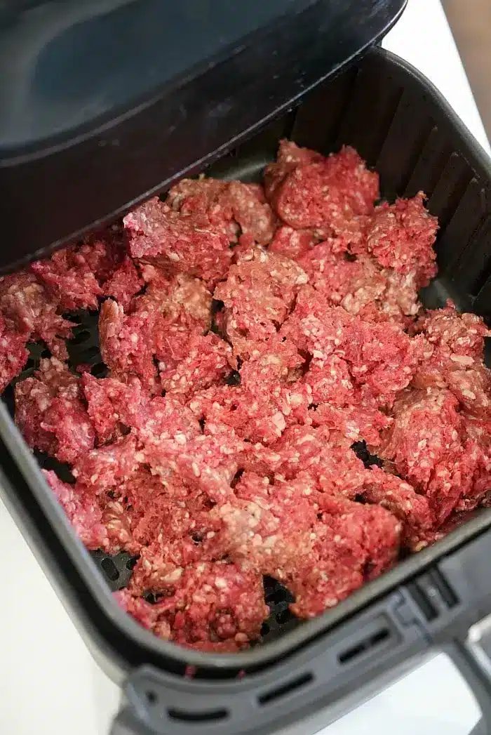 Ground beef is spread out in an air fryer basket, ready to be cooked. The air fryer lid is partially visible at the top of the image, solid for those exploring new air fryer beef recipes.
