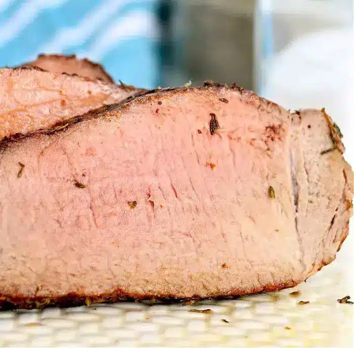 A close-up view of a thick, sliced piece of cooked meat with a pink interior and slightly charred outer edge, solid for air fryer beef recipes. The background is slightly blurred, showing a blue and white striped cloth and a surface.