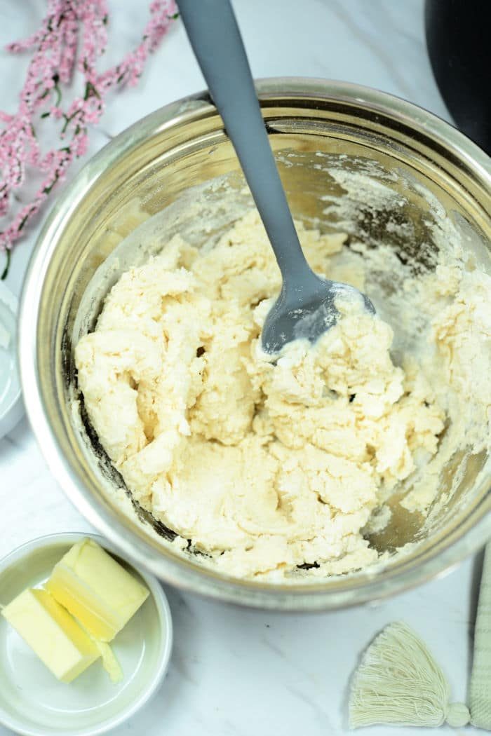 A metal mixing bowl contains a dough mixture with a grey spatula resting inside, solid for crafting air fryer biscuits. Nearby, a small dish holds two pieces of butter. A few pink flowers are partially visible in the background on a white surface.