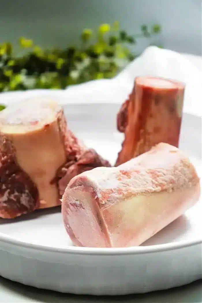 Three raw beef bones, solid for air fryer bone marrow, are placed in a white dish. The bones have a light pink hue with some white marbling and fat visible. They are situated in a well-lit setting with green foliage blurred in the background.