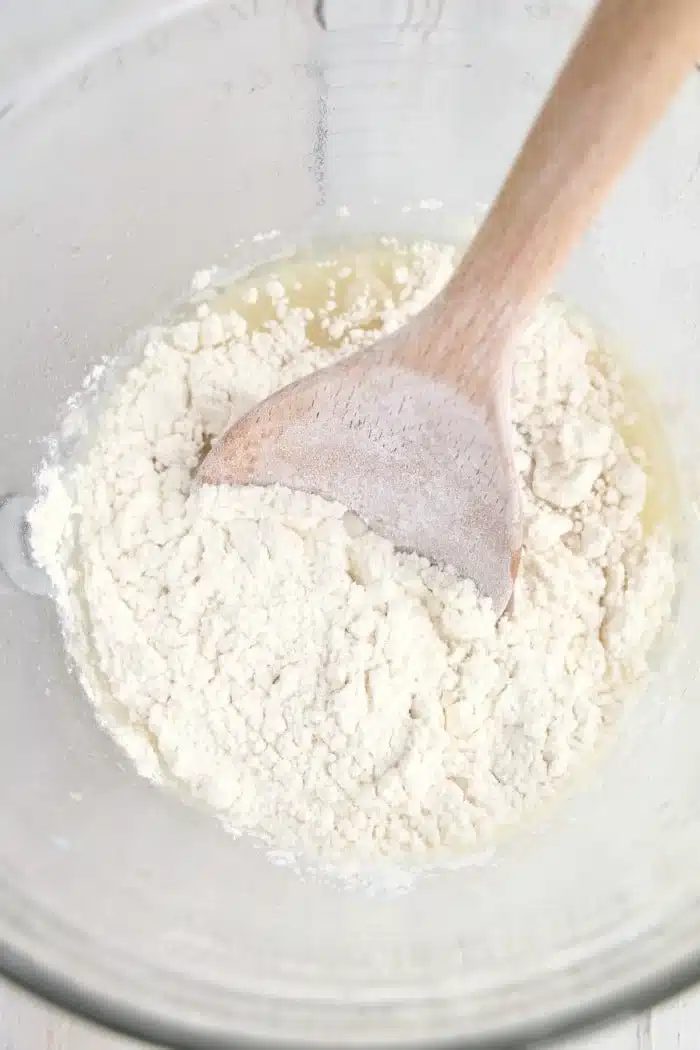 Wooden spoon mixing flour and liquid in a clear glass bowl, preparing dough for good air fryer bread.