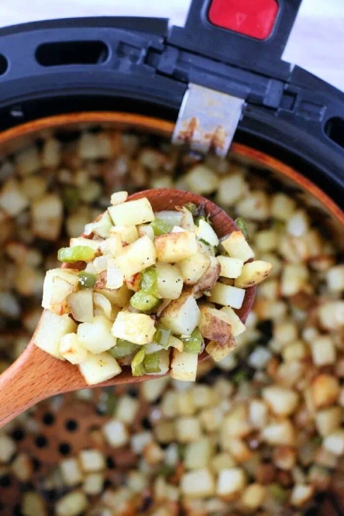 A wooden spoon holds diced potatoes and vegetables over an air fryer basket filled with air fryer breakfast potatoes.