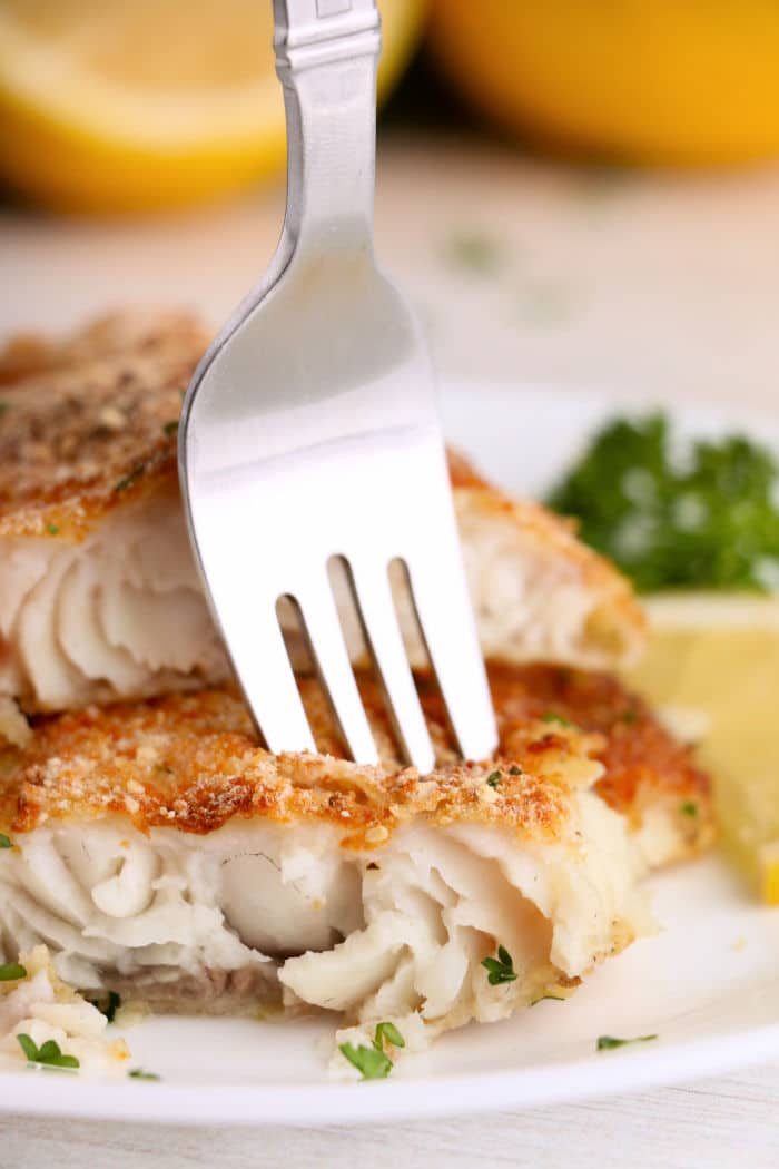 A fork cutting into a piece of crispy air fryer catfish, served with parsley and a lemon wedge on a white plate.