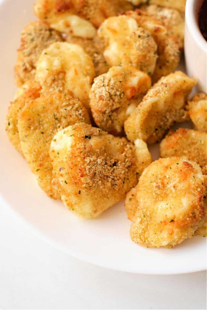 A plate of crispy, golden brown air fryer cheese curds, perfectly seasoned and mouth-wateringly good, with a small bowl of marinara sauce partially visible on the side.
