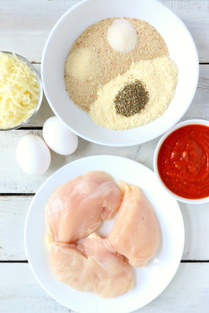 Bowls of ingredients on a white surface include raw chicken breasts, breadcrumbs with herbs and spices, marinara sauce, grated cheese, and two eggs—solid for crafting good air fryer chicken parm.