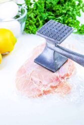 Raw chicken breast being tenderized with a metal mallet, surrounded by eggs, parsley, and a lemon. Wondering how long to bake chicken cutlets? This setup is solid for creating the tender base you'll need before popping them in the oven for good results.