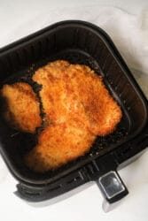Breaded chicken cutlets, reminiscent of air fryer chicken katsu, are cooking in an air fryer basket. The cutlets are golden brown and crispy, set against a black basket surface on a white background.