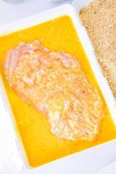 Raw chicken breast marinating in a yellow mixture, next to breadcrumbs in a white tray, waiting to become good baked cutlets. For perfectly cooked cutlets, consider how long to bake chicken cutlets at your desired temperature and enjoy a tender culinary delight.
