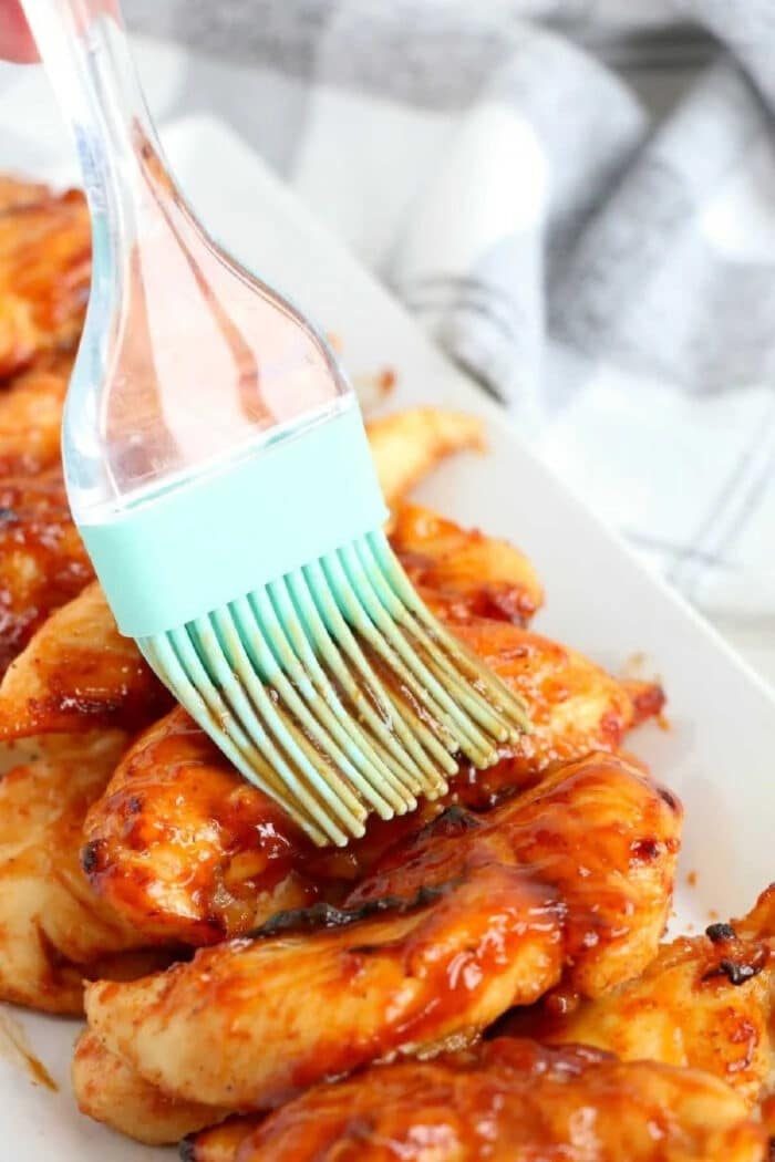A silicone brush applies savory barbecue sauce to juicy air fryer chicken tenders with no breading, arranged on a white plate.
