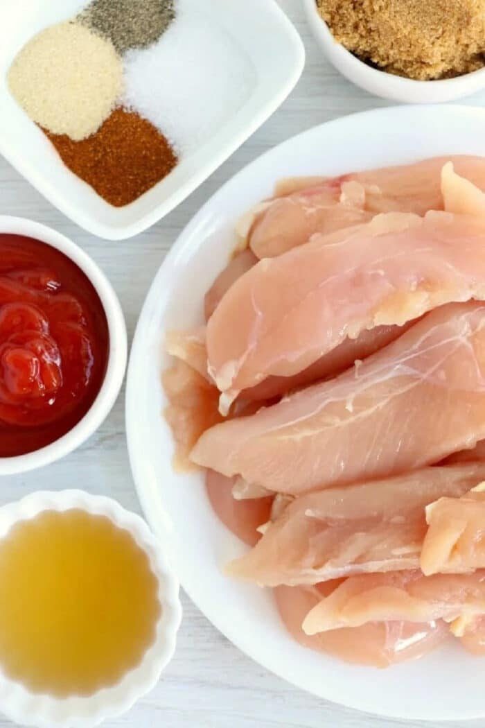 Raw chicken strips on a plate, ready to become good air fryer chicken tenders with no breading, are surrounded by bowls of ketchup, spices, brown sugar, and a yellow liquid for flavor.