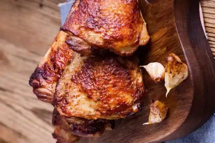 air fried bone-in chicken thighs with crispy, browned skin are arranged on a wooden cutting board. Garlic cloves, also roasted, are placed next to the chicken pieces. The background features a wooden surface with a rustic appearance.