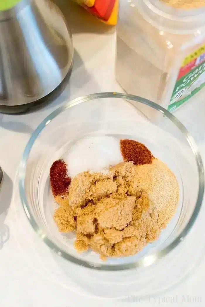 A glass bowl containing various seasonings, including brown sugar, garlic powder, paprika, and salt. A metal object and a container of spices are partially visible in the background on the white surface—solid for flavoring bone-in air fryer chicken thighs.