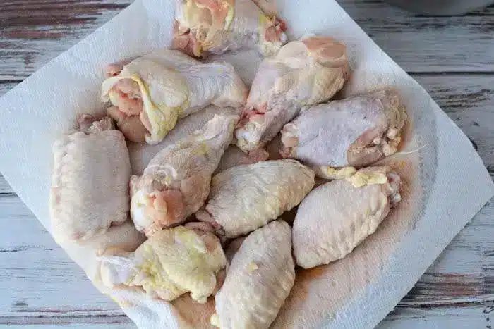 A collection of raw chicken wings and drumsticks are laid out on a white paper towel on a rustic, white wooden table. The fresh chicken pieces are ready for preparation or cooking, and would be solid as air fryer chicken wings when seasoned with a touch of baking powder.