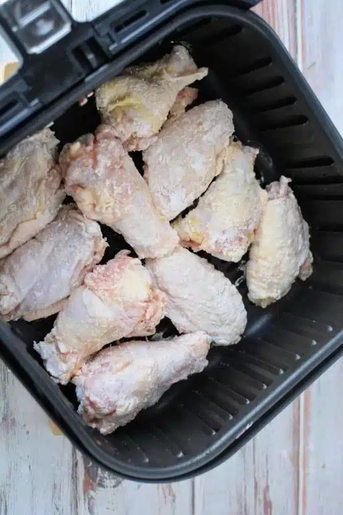 A close-up of raw, seasoned chicken wings dusted with baking powder, placed inside a black air fryer basket and ready for cooking. The countertop beneath reveals a light-colored surface with a faint wood grain pattern.