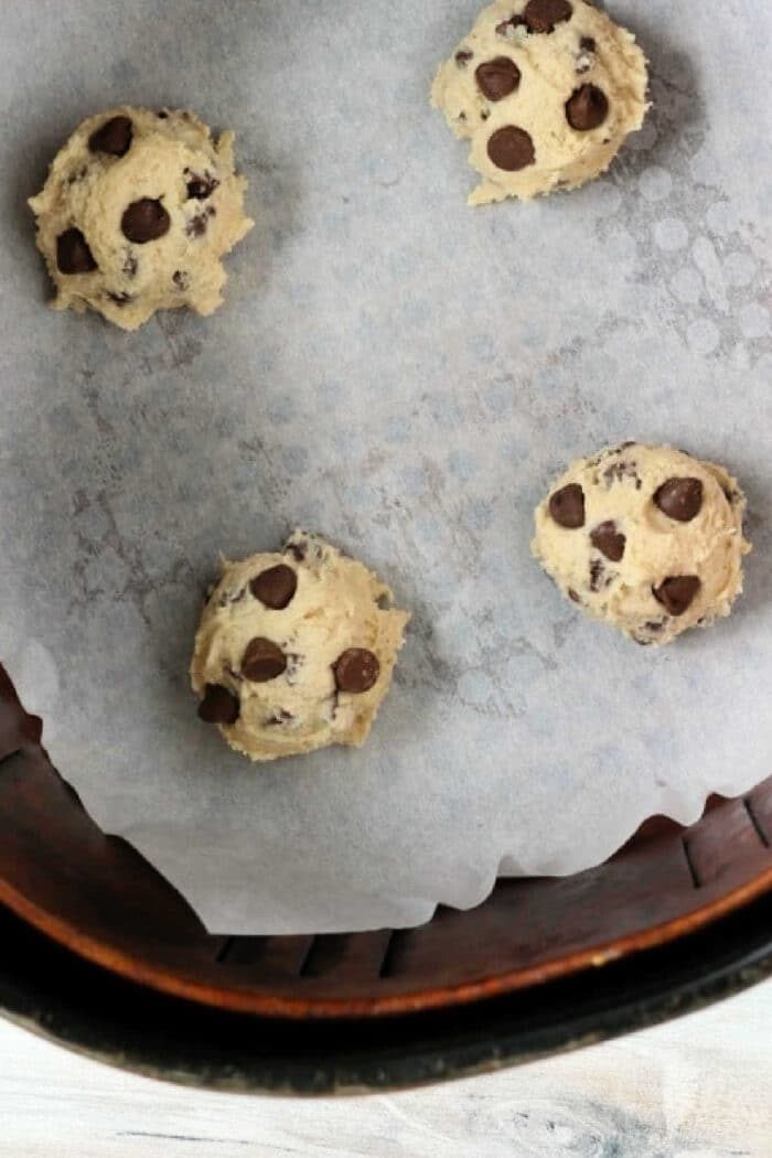 Four air fryer cookie dough balls arranged on a parchment-lined baking sheet. The dough balls are spaced apart, ready for baking. The bottom edge of a circular container is visible at the bottom of the image.