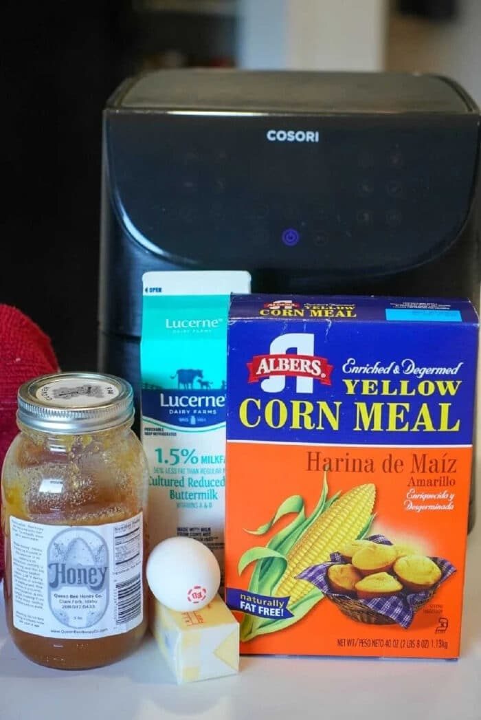 A carton of buttermilk, cornmeal box, honey jar, egg, and butter stick are arranged in front of an air fryer—solid ingredients for making good air fryer cornbread.
