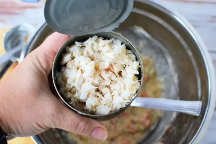 A hand is holding an open can of crab meat over a stainless steel mixing bowl. A spoon is visible inside the bowl, which contains a mixture of chopped vegetables and other ingredients for Air Fryer Crab Cakes.