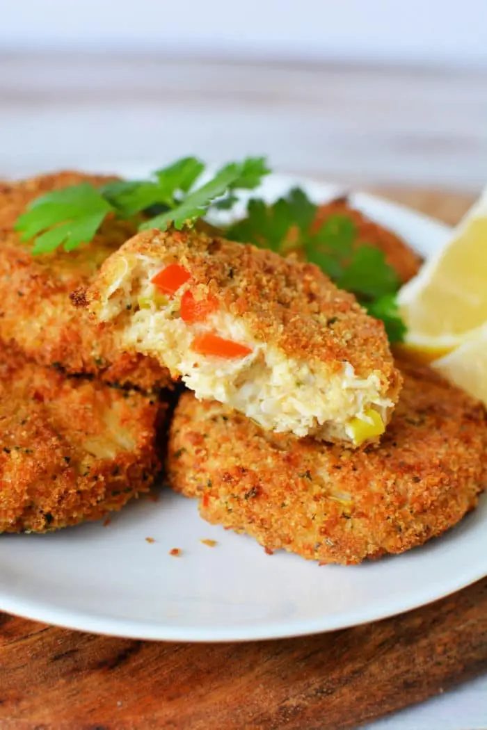 A white plate holding a stack of golden-brown, air fryer crab cakes garnished with fresh parsley. One crab cake on top has a bite taken out, revealing a mixture of crab meat and diced vegetables inside. A lemon wedge is placed beside the stack.