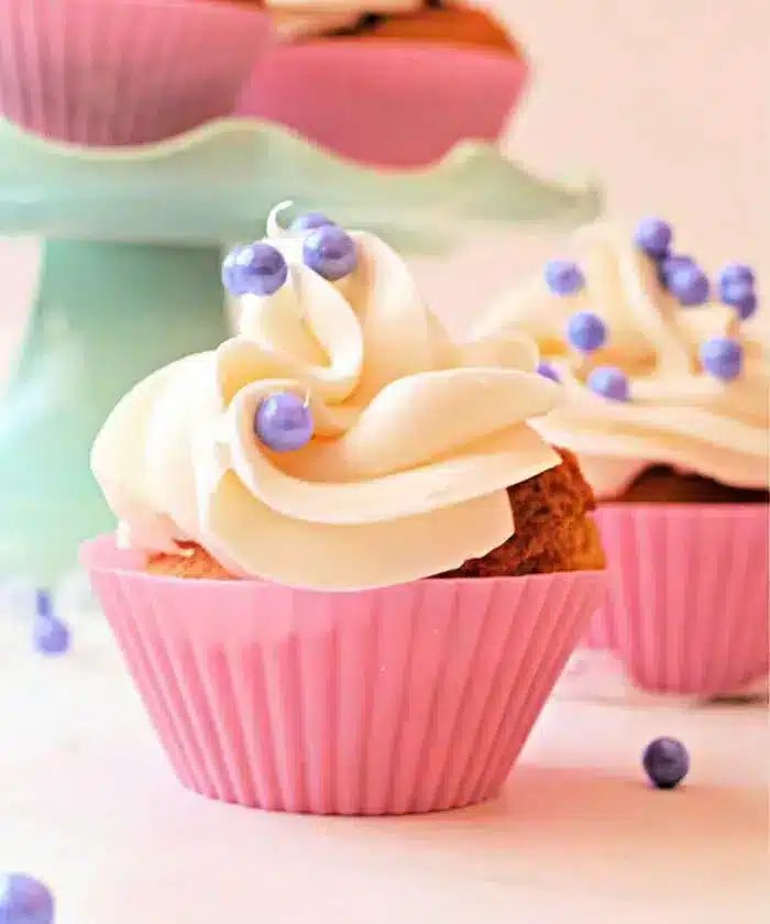 A cupcake with white frosting and purple candy pearls, made expertly in an air fryer, sits in a pink cupcake liner.