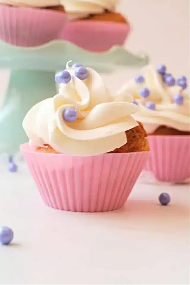 A close-up of air fryer cupcakes in pink silicone wrappers, topped with white swirled frosting and decorated with small purple pearl-like sprinkles.