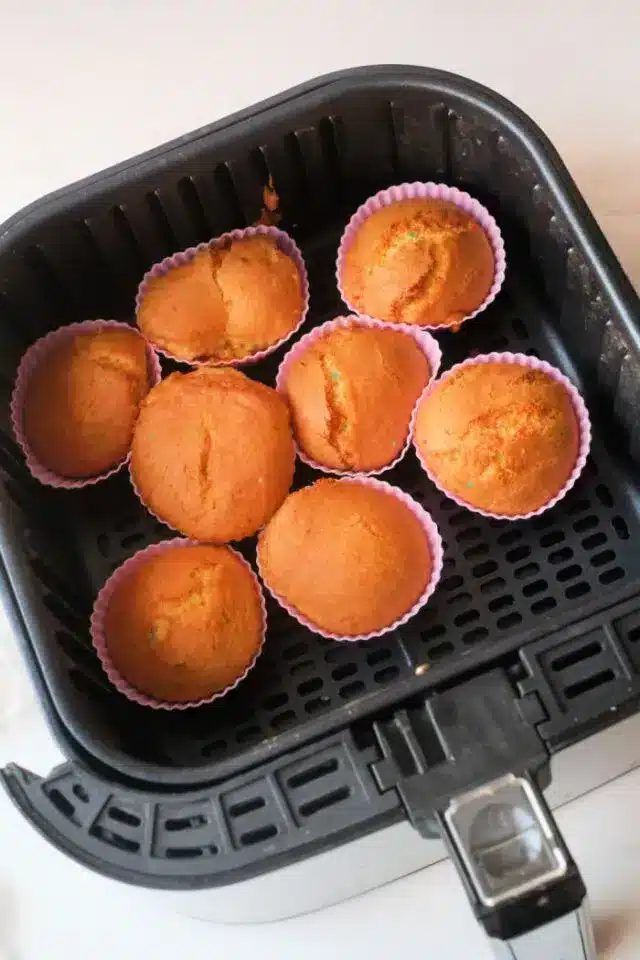 Seven freshly baked air fryer cupcakes in pink silicone cups are arranged in the basket of a black air fryer. The cupcakes are golden-brown and appear to have a slightly cracked surface. The air fryer is positioned on a light-colored countertop.