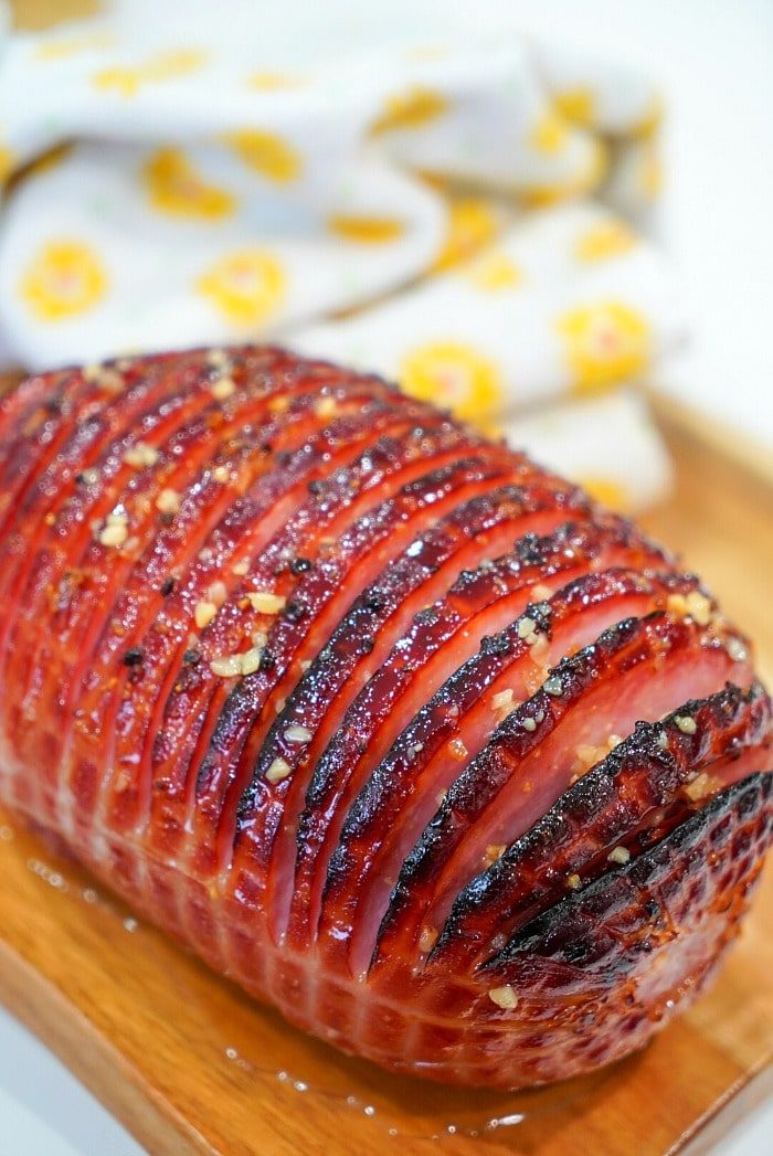 Close-up of a glazed, roasted air fryer ham with scored lines on a wooden board, with a patterned cloth in the background.