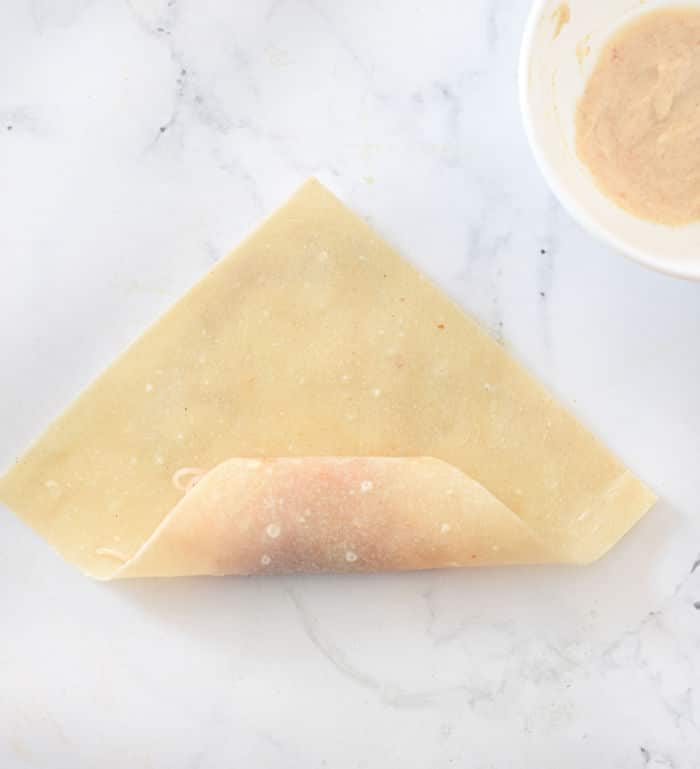 A triangular egg roll dough being folded on a marble surface, with homemade egg roll wrappers nearby and a bowl of filling ready to be savored.