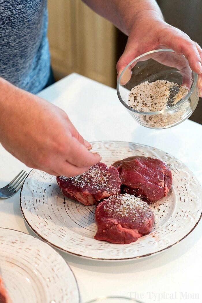 A person seasons three raw pieces of steak with a mix of salt and pepper from a small glass bowl, preparing them perfectly for the grill. The steaks rest on a white plate with decorative patterns, ready to be transformed by smoky flavor. A fork is visible beside the plate on a white countertop.
