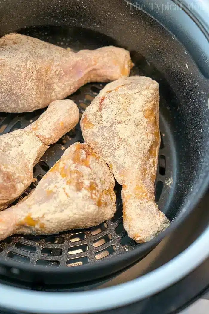 Close-up of four raw, breaded chicken drumsticks placed in the basket of an air fryer, ready to be cooked. The chicken is lightly coated with a flour mixture, solid for an easy air fryer fried chicken recipe. The air fryer basket's black mesh is visible underneath the drumsticks.