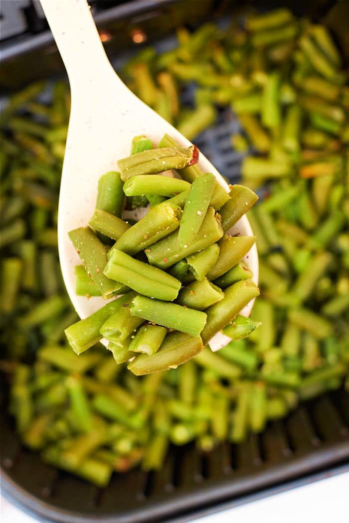 Close-up of a spoon holding air fried green beans over a pan filled with more green beans, showcasing the vibrant freshness often missing in air fryer frozen green beans.