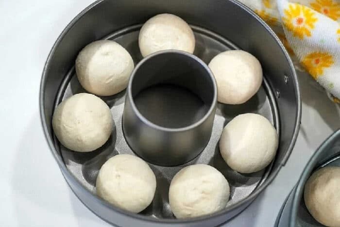 Eight dough balls, solid for making Frozen Rolls in Air Fryer, are arranged in a ring mold pan and ready for baking on a white surface.