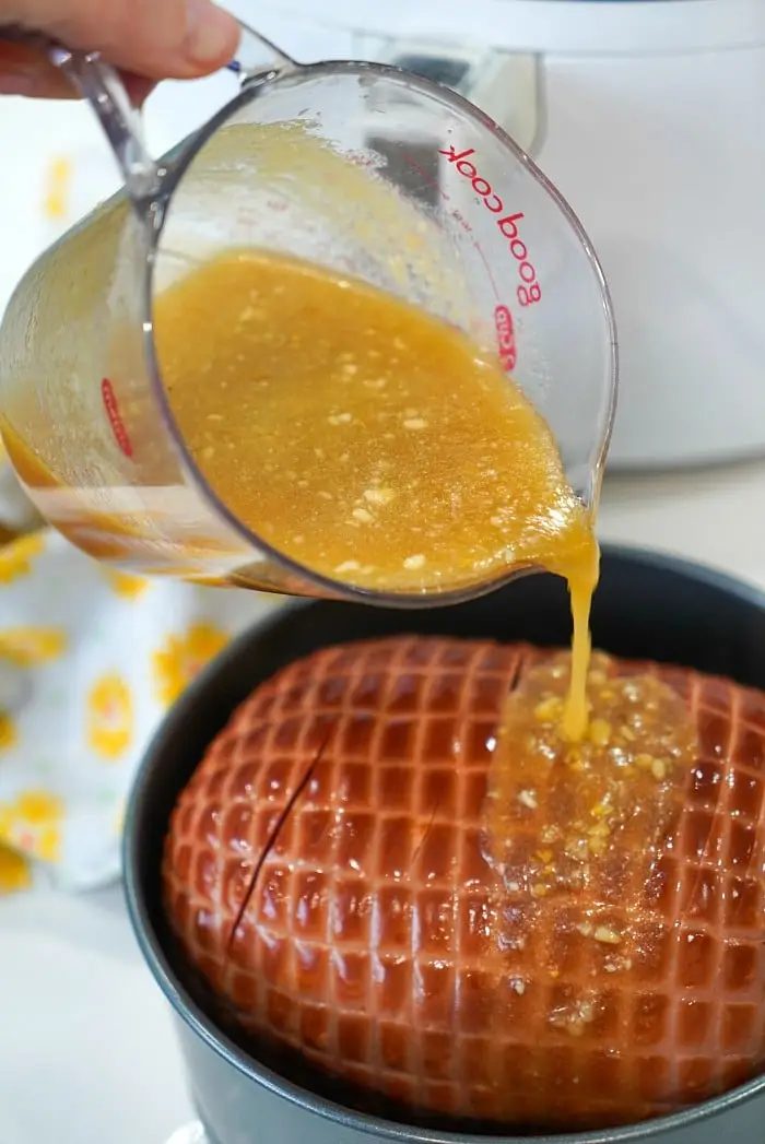 Close-up of a hand pouring a pineapple glaze from a glass measuring cup onto an air fryer ham in a black pot. In the background, you can see a white appliance and a yellow and white patterned cloth.