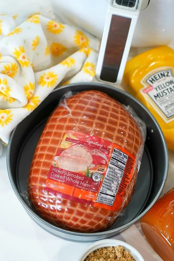 A round pan containing a packaged hickory-smoked cooked whole ham sits on a counter, ready to be transformed into a good air fryer ham. Surrounding items include a tub of yellow mustard, a small container of brown sugar, a meat thermometer, and a cloth with a yellow floral pattern.