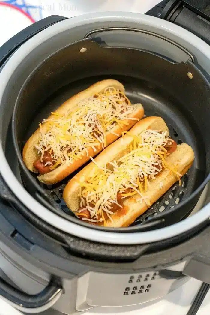 Two air fryer hot dogs with grated cheese on top sit in a basket, nestled in their buns side by side. The air fryer lid is visible at the edge of the image, capturing a solid meal prep moment.
