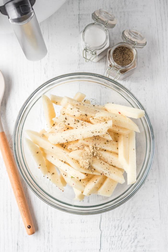 A glass bowl holds air fryer jicama fries, seasoned to perfection with spices and herbs, while a wooden spoon and spice jars rest nearby.