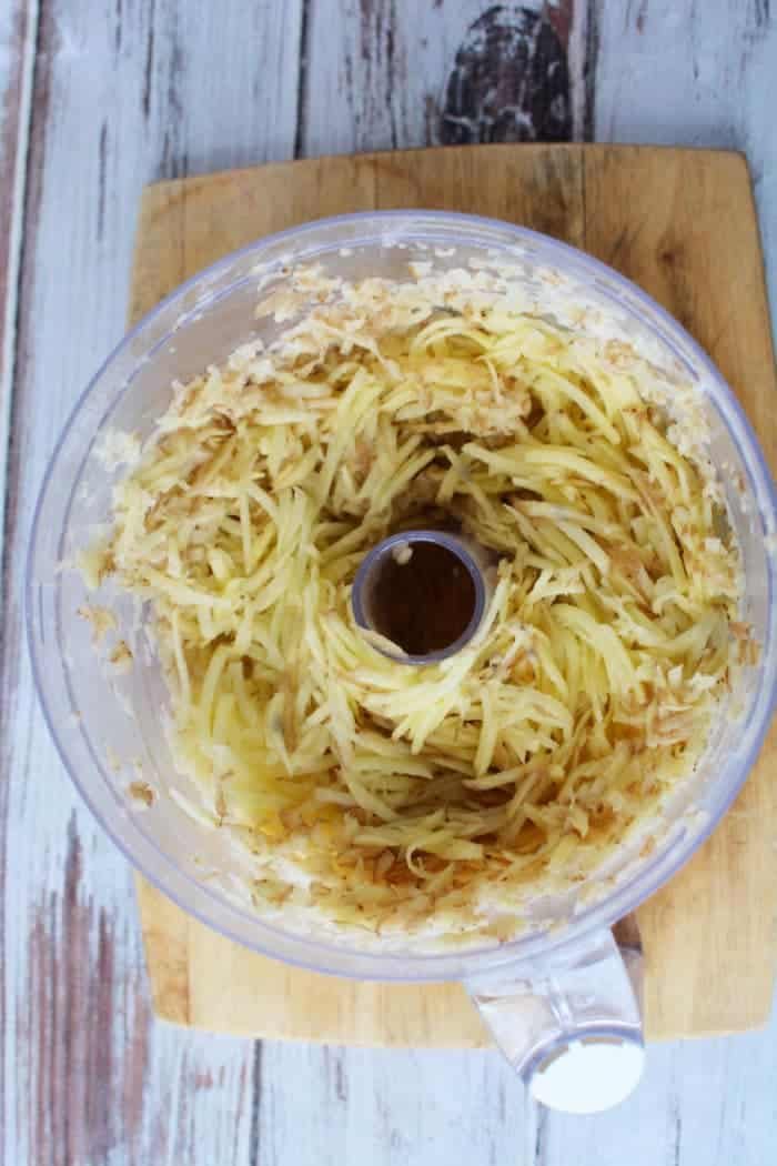 Shredded potatoes in a food processor bowl on a wooden cutting board, ready to be transformed into crispy Air Fryer Latkes.