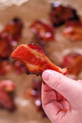 A hand holding a crispy piece of Air Fryer Maple Candied Bacon, with more maple-glazed bacon pieces blurred in the background.